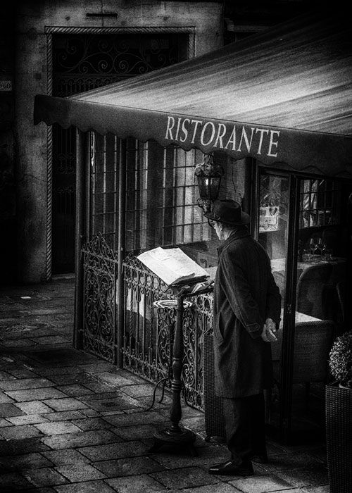 Ein Mann mit Hut und Mantel steht vor einem Restaurant und wartet auf Kundschaft. Street Photography aus Venedig in schwarz weiß.