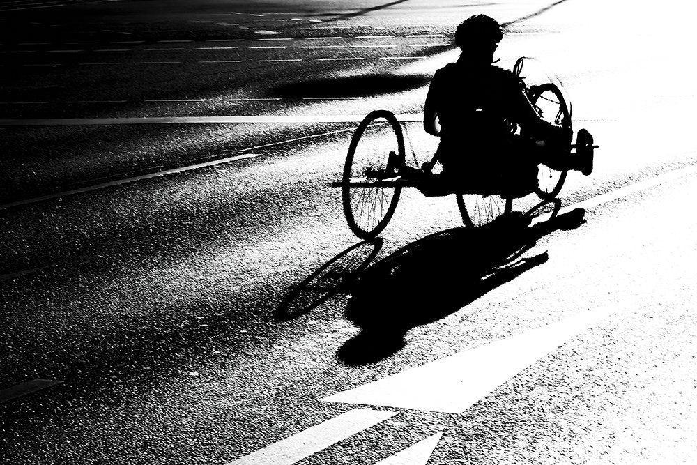 Ein Rollstuhlfahrer fährt auf einer Straße in Richtung Sonnenlicht. Er wirft einen großen Schatten. Abstrakte Straßenfotografie aus Berlin in schwarz weiß
