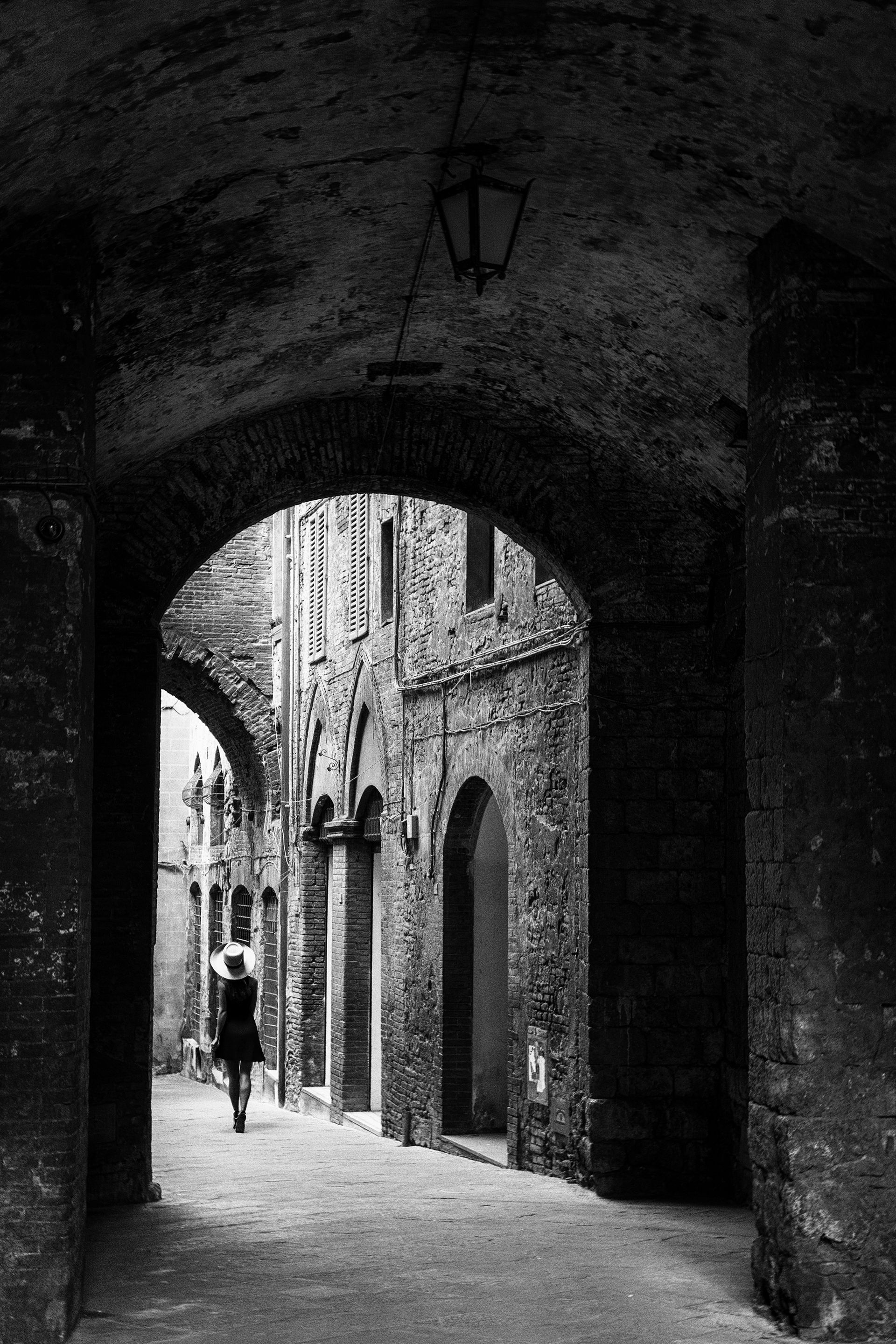Schwarz weiß Fotografie einer Frau mit Kleid und Hut in einer Gasse der Altstadt von Siena Toskana