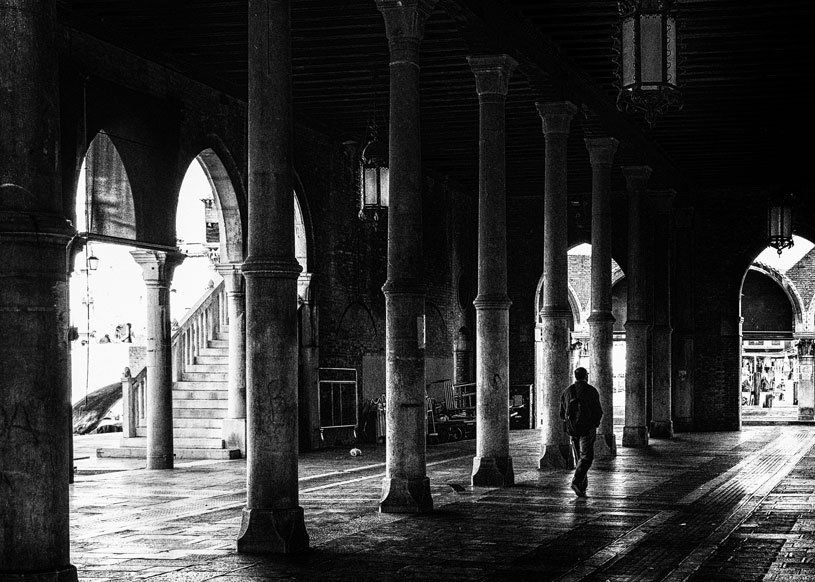 Ein Mann geht allein durch die leere Säulenhalle des Fischmarktes in Venedig. Straßenfotografie aus Italien in schwarz weiß.