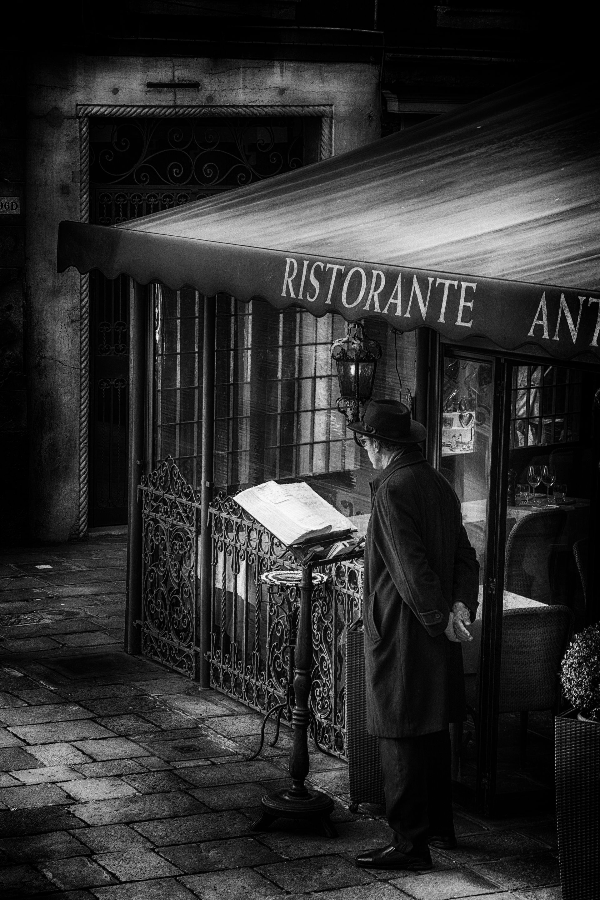 Schwarz weiß Fotografie eines Mannes mit Mantel und Hut vor einem Restaurant in Venedig. 