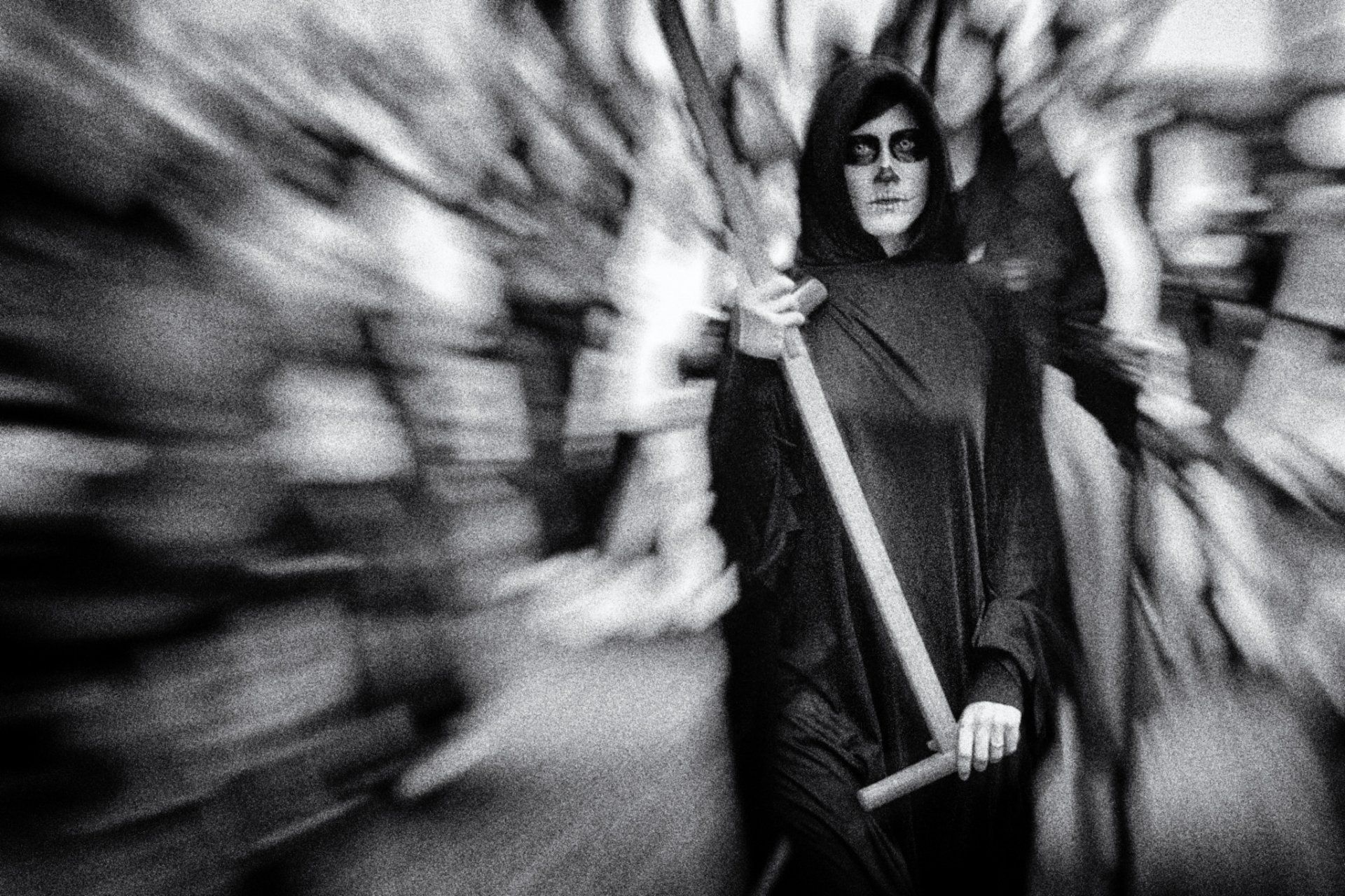 Eine junge Frau als Tod verkleidet mit Sense in der Hand bei einem Umzug auf der Straße. Abstrakte Straßenfotografie in schwarz weiß