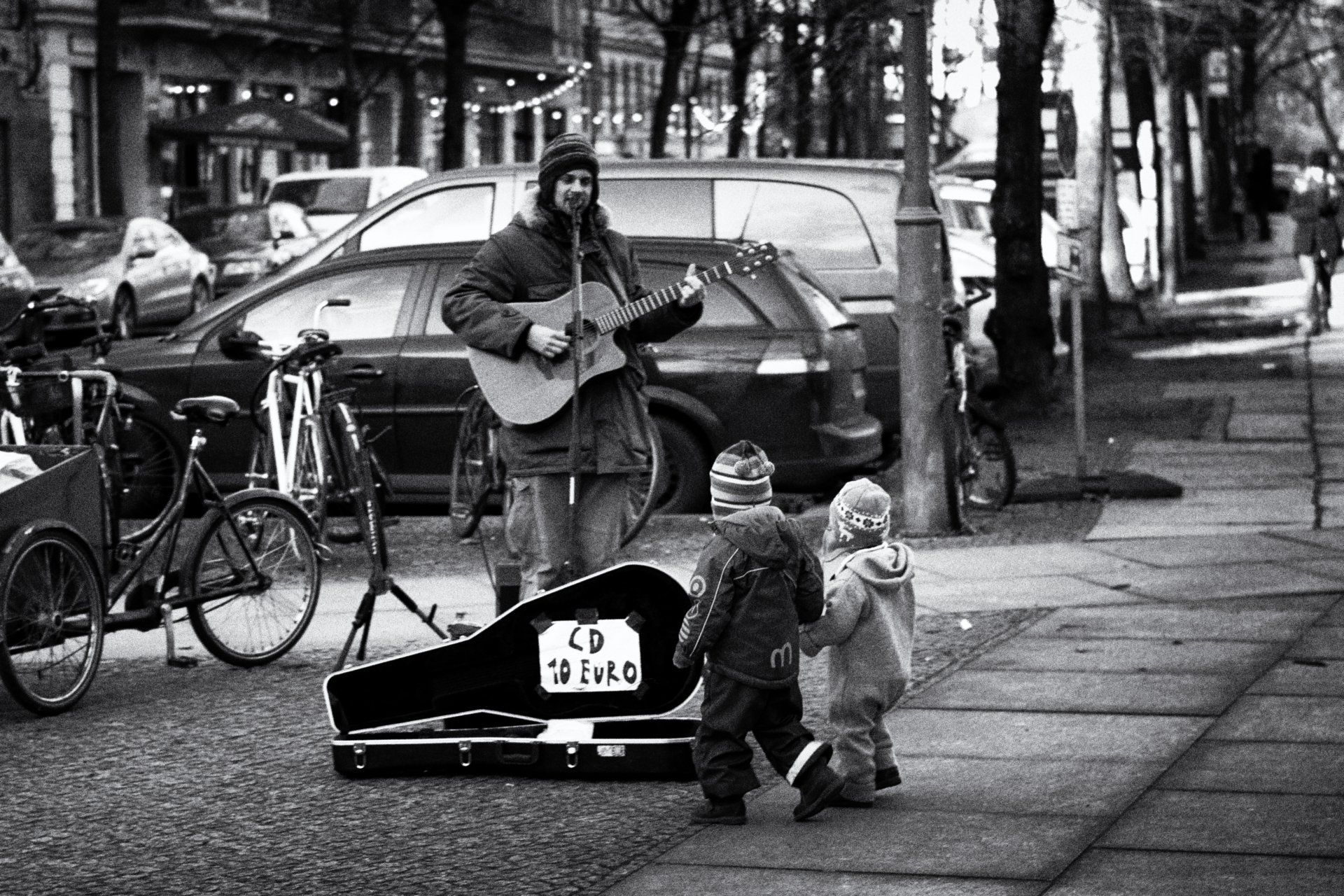 Ein Straßenmusiker spielt Gitarre im Prenzlauer Berg. Zwei kleine Kinder stehen Hand in Hand vor dem Gitarristen und hören ihm zu. Street photography aus Berlin in schwarz weiß.