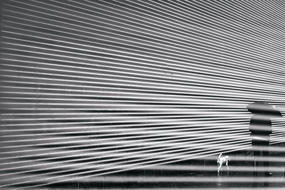 Eine Frau mit Regenschirm geht mit ihrem Hund im verregnten Berlin spazieren. Abstrakte Straßenfotografie aus Berlin in schwarz weiß.