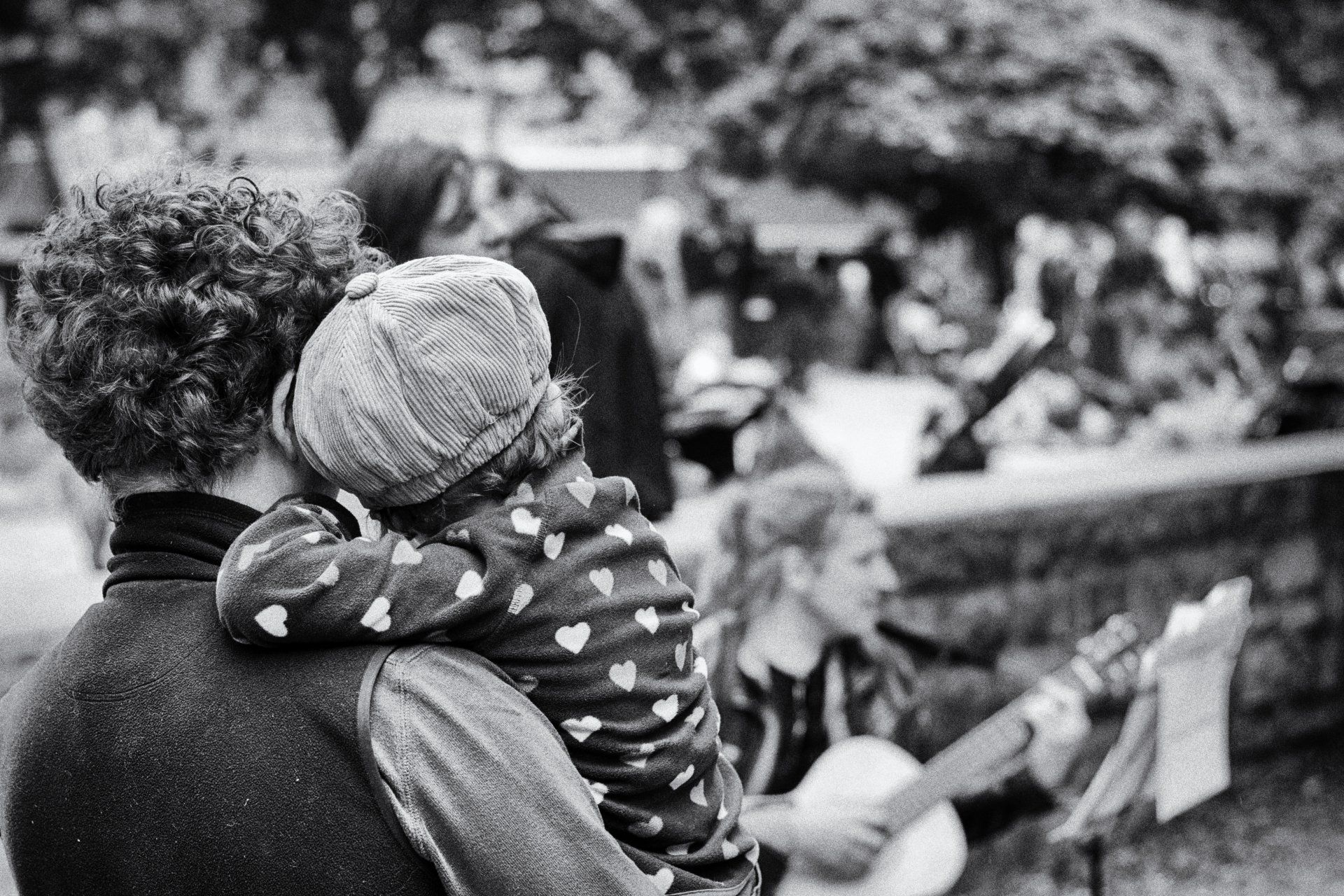 Ein Vater hält sein Kind in den Armene. Die beiden hören innig einem Straßenmusiker auf dem Kollwitzplatz zu. Street photography aus Berlin in schwarz weiß.