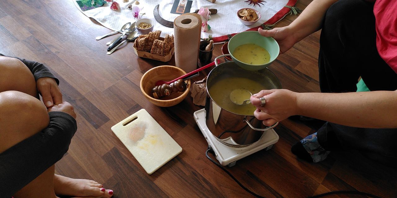 Topf mit Suppe, Frau gibt mit Suppenkelle Suppe in eine Schale, zweite Frau sitzt gegenüber