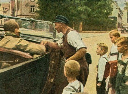 1930's man and children dressed in German lederhosen. The man is talking to a person in a car.