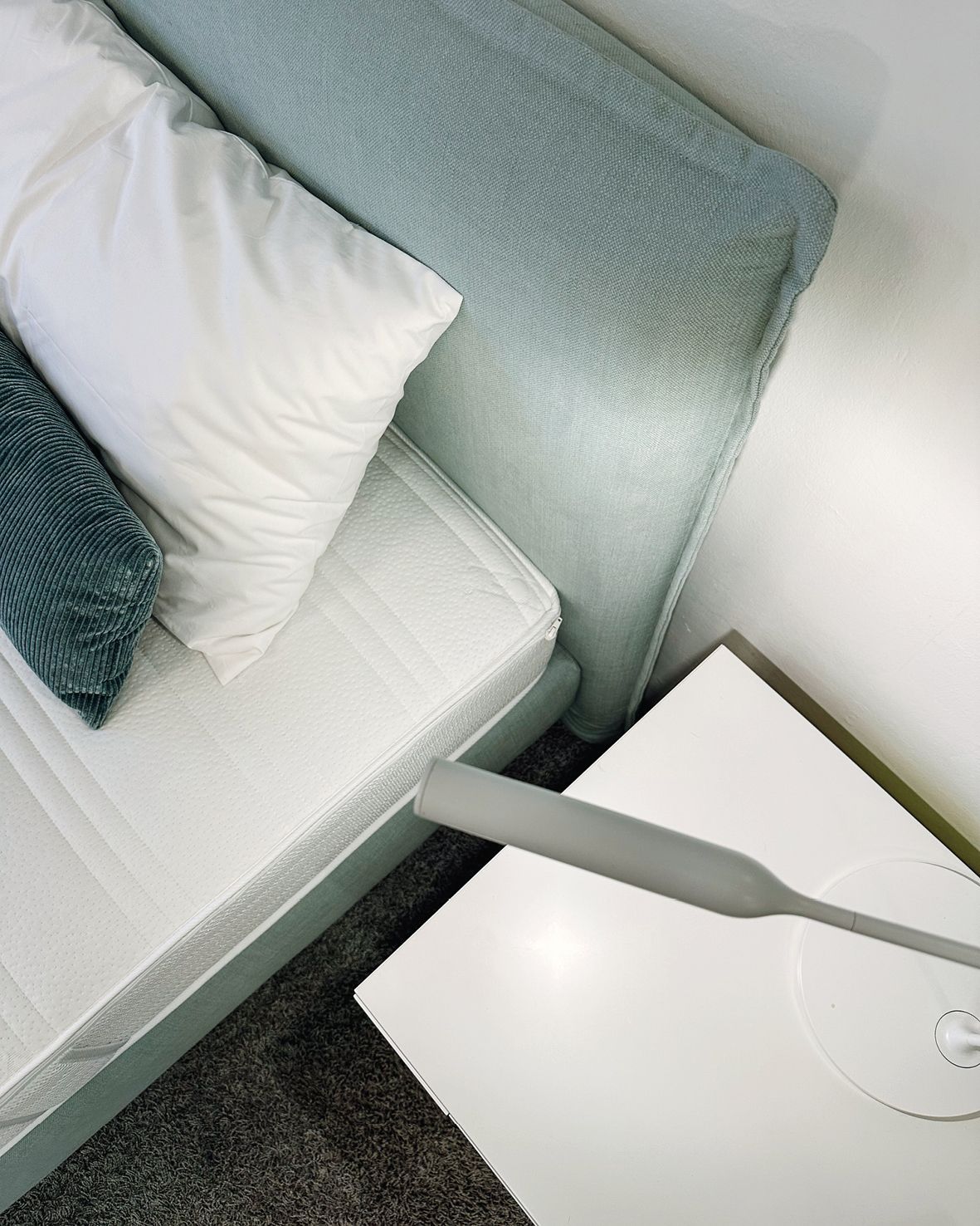Detail eines Boxspringbettes, oben die Matratze, Kissen und der Ausschnitt des Kopfteils, daneben ein weißer Nachttisch und eine schmale Tischleuchte