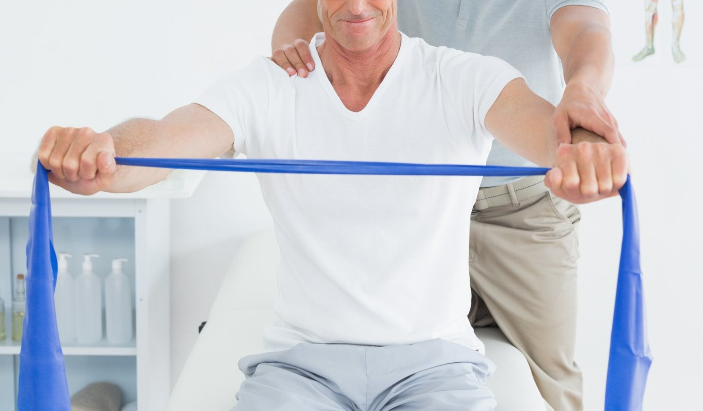 Eine Person sitzt und zieht ein Therapieband auseinander, ein Therapeut hat seine Hände auf das linke Handgelenk und auf die rechte Schulter gelegt