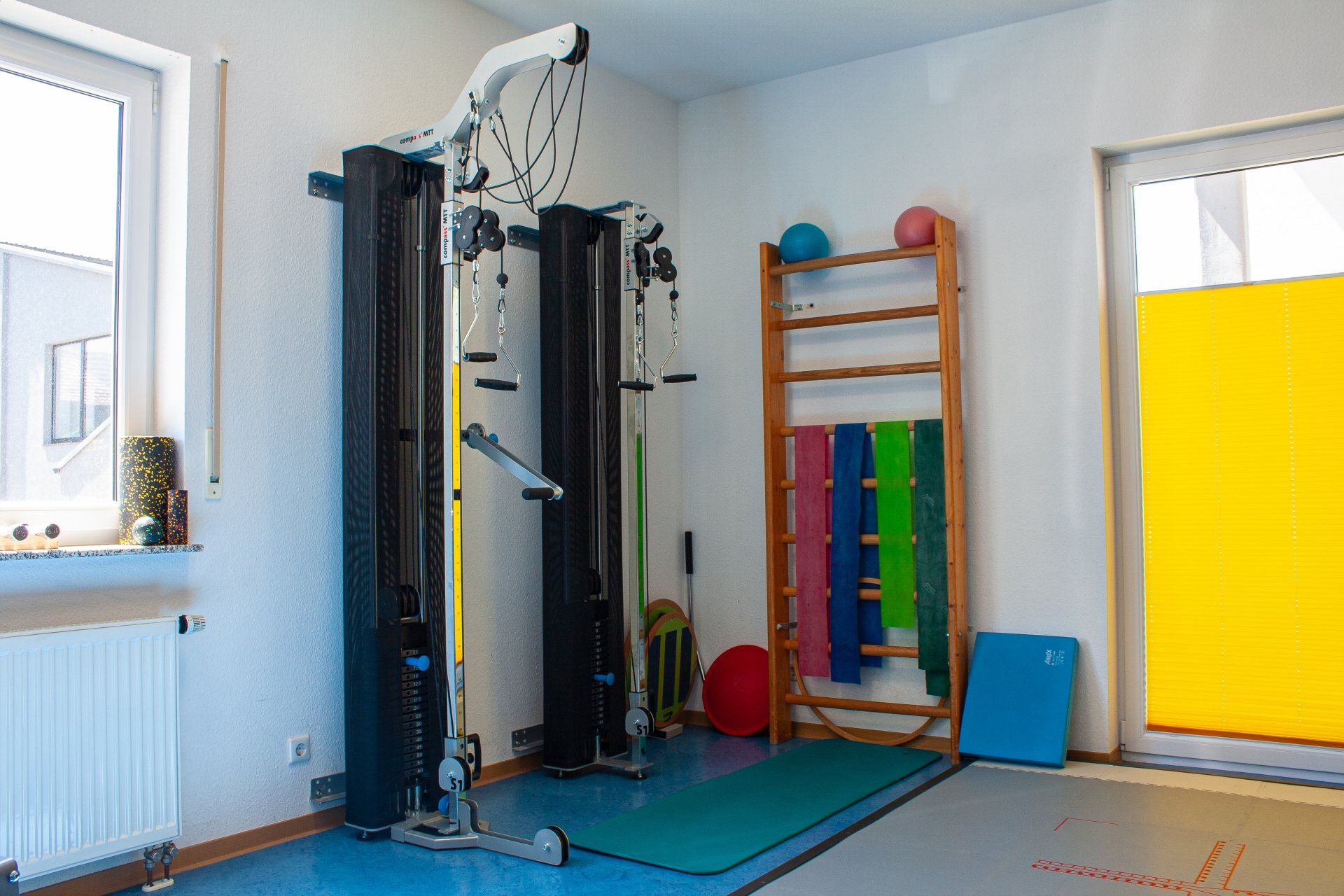 Ausschnitt aus einem Trainingsraum. Links befindet sich ein Seilzug, in der Mitte eine Sprossenwand mit verschiedenen Therapiebändern.