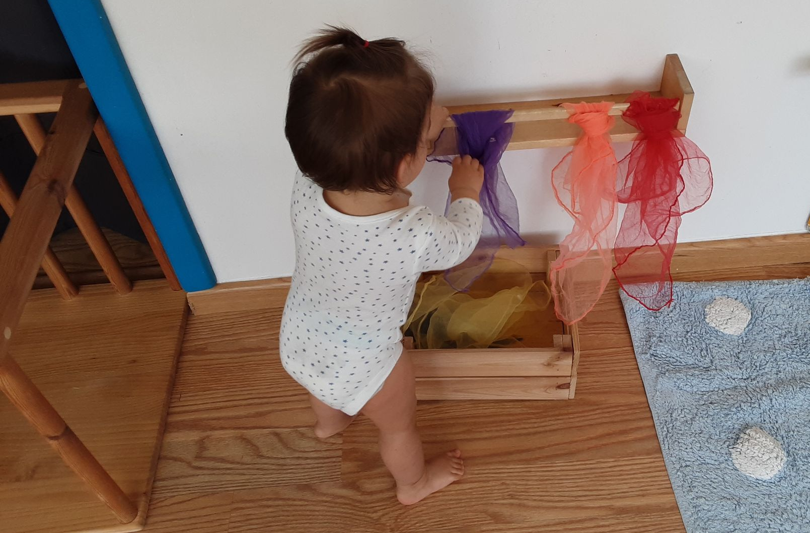 Bebé de pie agarrado a una barra con pañuelos de colores en el ambiente de Nido de Montessori Boadilla