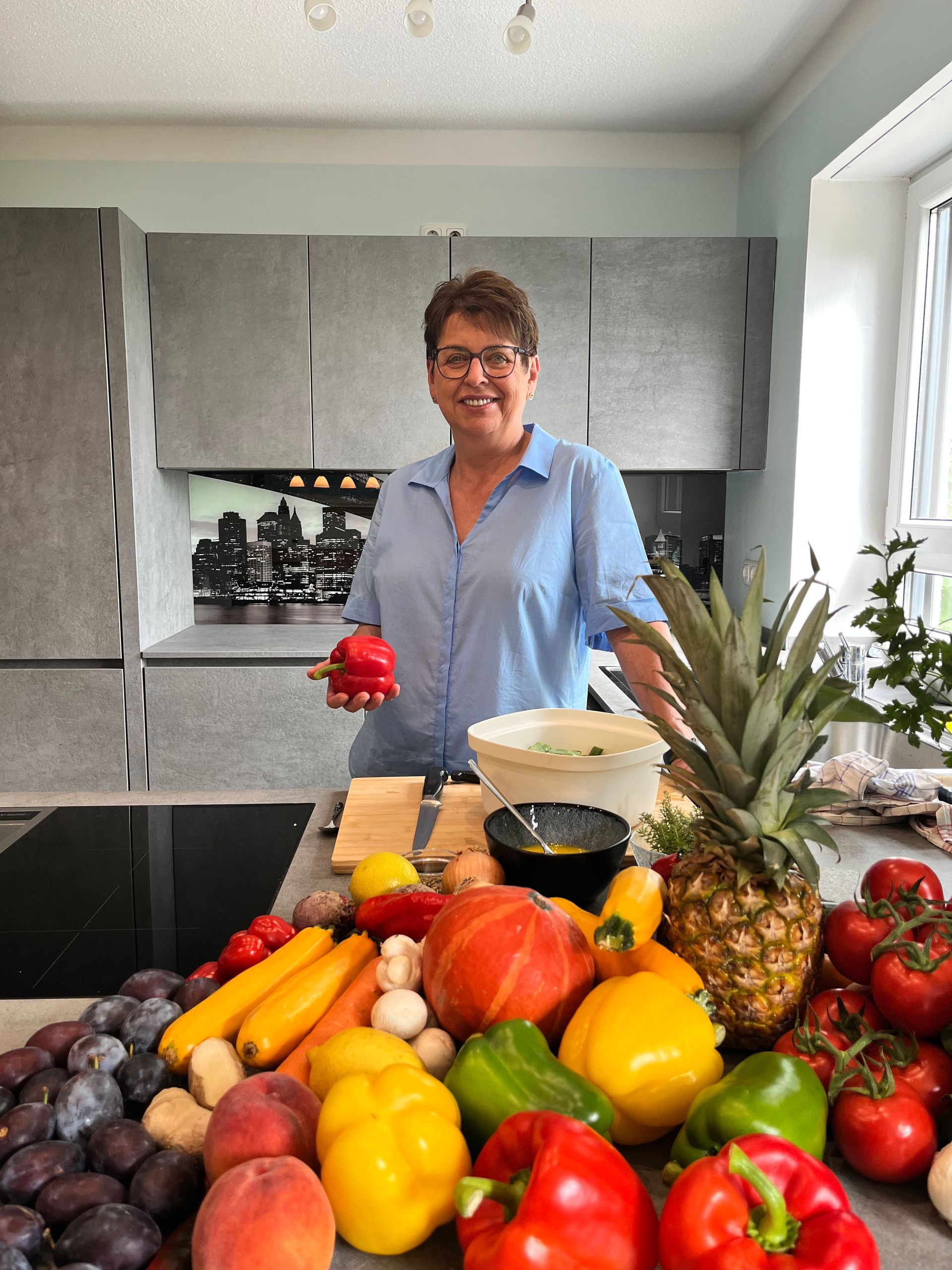 Elke Landisch, Ernährungsberaterin, Ayurvedaköchin und Köchin aus Landshut mit einer Paprika in der Hand hinter einer großen Auswahl an Obst und Gemüse.