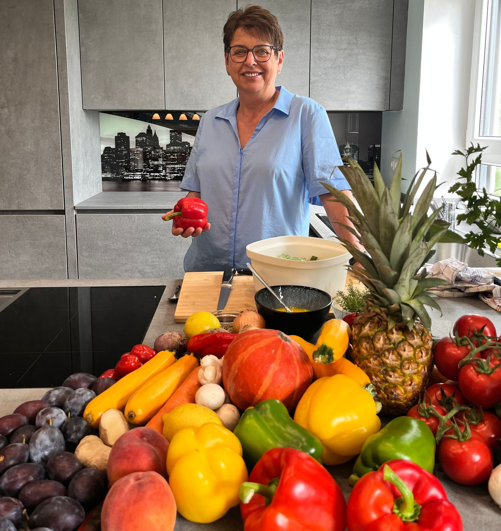 Elke Landisch, Ernährungsberaterin, Ayurvedaköchin und Köchin aus Landshut mit einer Paprika in der Hand hinter einer großen Auswahl an Obst und Gemüse.