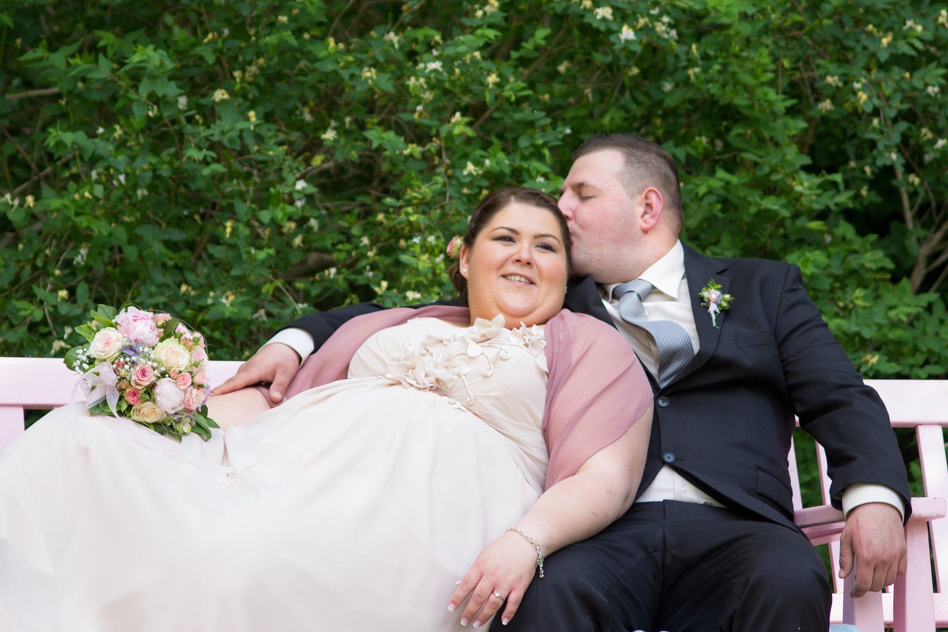 Hochzeitspaar in Großaufnahme auf Bank sitzend, er gibt ihr einen Kuss auf die Stirn