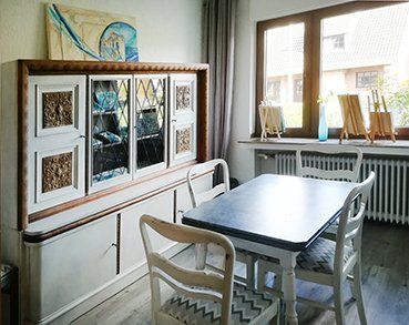 Ansicht vom Wohnzimmer ins Esszimmer. Der Esszimmertisch und die 4 Stühle stehen vor dem Großen Fenster das zum Vorgarten und dahinter zur ruhigen Kreuzstraße weißt. Links davon steht ein geräumiger Vitrinen Schrank. Alle Möbel sind im Vintage-Style und in weiß-grauen Farbschattierungen gehalten, ergänzt durch einige dunkeln holzfarbigen Akzenten.