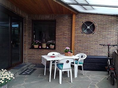 Große überdachte Terrasse mit Natursteinplatten zugänglich. Darauf eine Sitzgruppe mit einem Rechteckigen weißen Tisch und weißen Stühlen. Geradeaus blickt auf ein Fenster zum Wohnzimmer daneben ist noch ein Bullaugenfenster, das zum Treppenhaus nach oben gehört. Darunter steht eine Polsterbox zur Unterbringung von Polstern und diversen Accessoires. Links ist die Terassentür, zugänglich über eine Stufe. Es stehen diverse Blumen auf der Terrasse am rechten Bildrand ist ein Fahrrad untergestellt.
