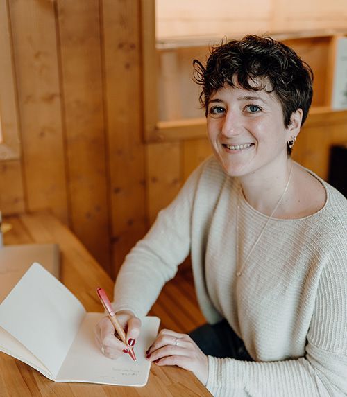 Steffi Müsse sitzt mit einem hellen Pullover an einem Tisch und schreibt in ein Notizbuch.