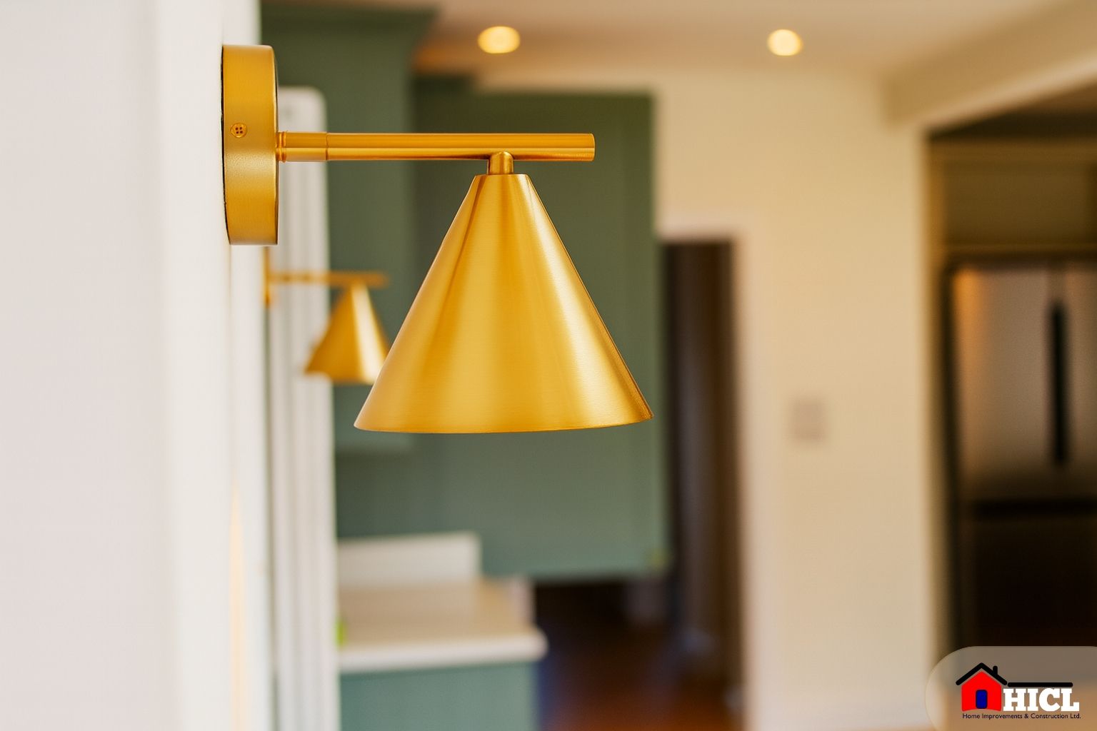 A close-up image of a brushed brass cone-shaped wall sconce, mounted on a white wall in a contemporary kitchen space, with soft ambient lighting and green cabinetry in the blurred background.
