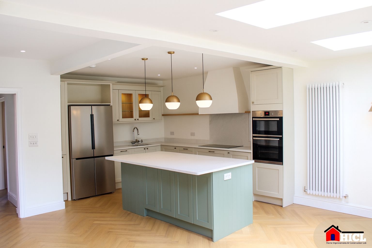 A high-resolution photograph showcases a spacious, modern kitchen with a sage green island, quartz countertops, brass pendant lighting, and herringbone wooden flooring, illuminated by overhead skylights.