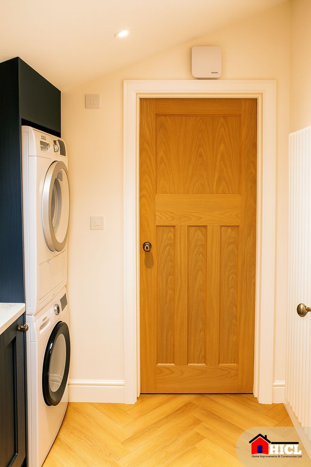 A compact and neatly designed utility room featuring a stacked washing machine and dryer, navy cabinetry with a white worktop, wooden herringbone flooring, and a natural oak door under a sloped ceiling.