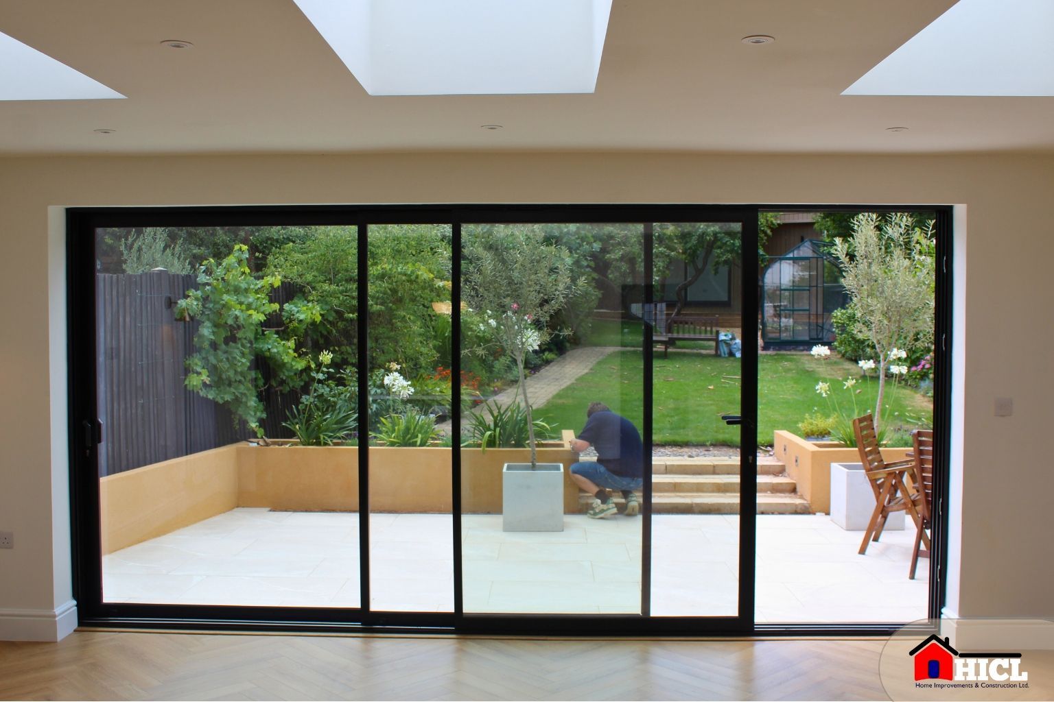 A bright, high-resolution image showing large sliding glass doors opening onto a landscaped garden with raised beds, outdoor seating, and a small greenhouse, viewed from a room with herringbone flooring and skylights.