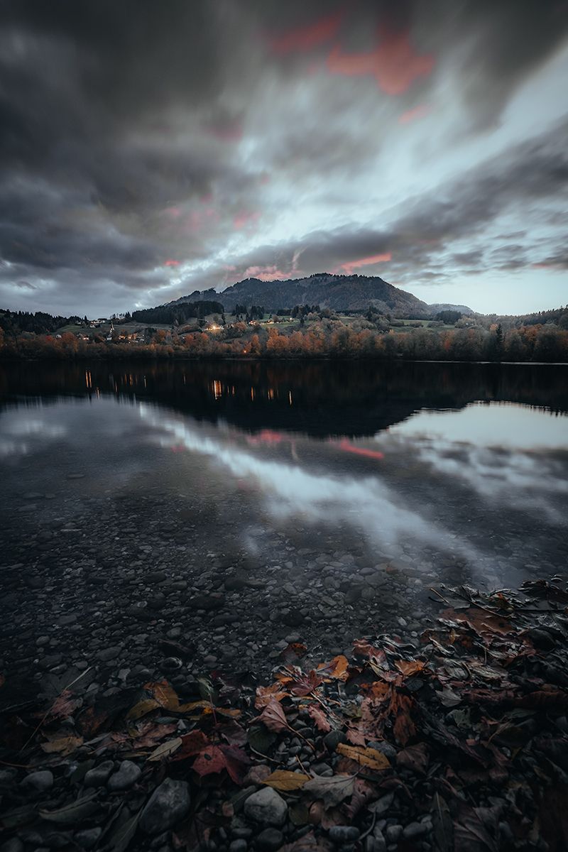 Allgäuer See zur blauen Stunde mit Spiegelung der Wolken im Wasser und herbstfarbenen Blättern im Vordergrund, Bayern