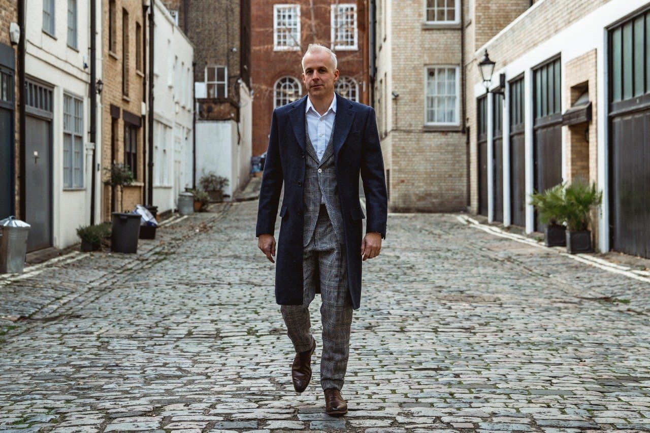 A gentleman wears a two-piece grey striped suit with a blue woollen fitted coat, all handmade by Adam Davies of Adam James Bespoke tailoring
