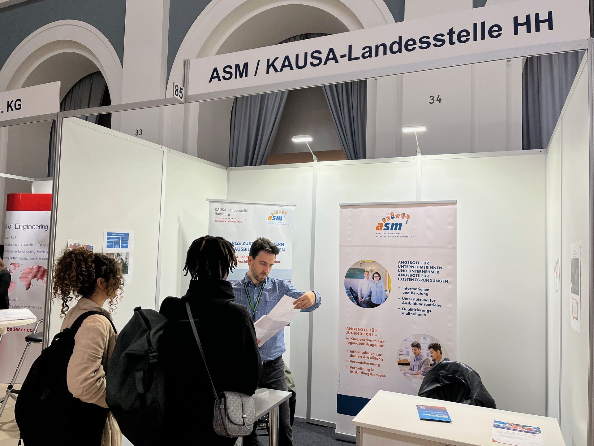 Am Stand der KAUSA-Landesstelle Hamburg auf der Lehrstellenbörse berät ein Mitarbeiter zwei Ratsuchende.