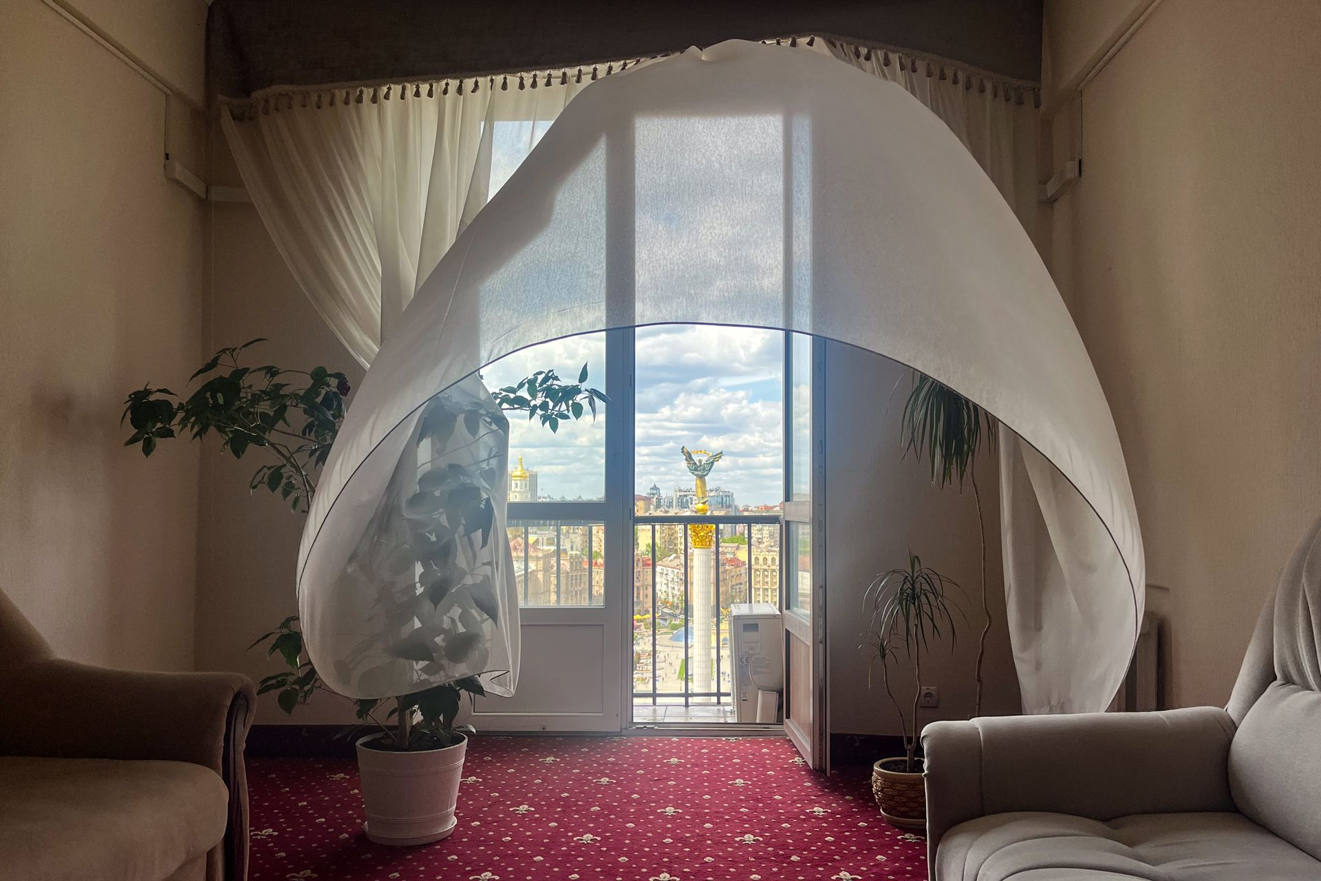 Curtain billowing in Kyiv hotel room with view of Independence Square and golden statue, spring 2025