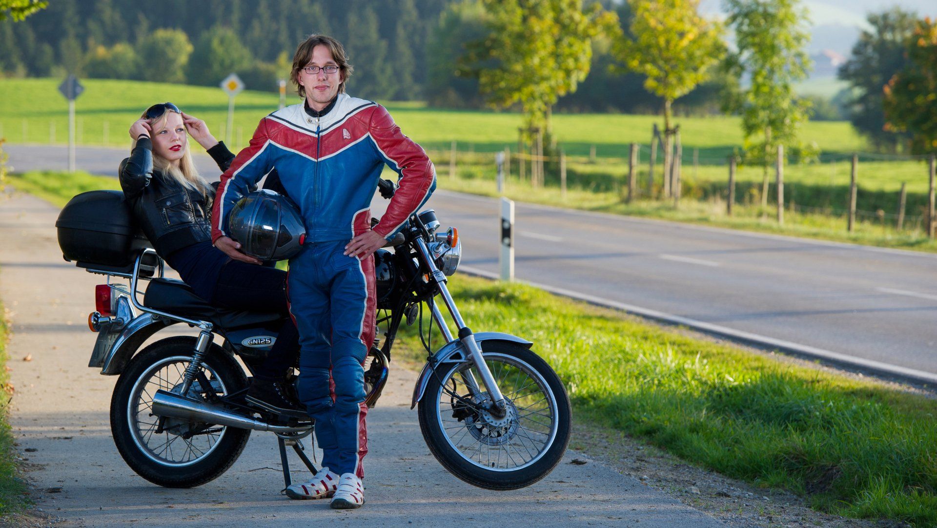 Auf diesem Bild ist ein Mann namens Herr Dressler zu sehen, der einen Motorrad Anzug trägt und einen Motorrad Helm in der Hand hält. Im Hintergrund befindet sich eine Frau, die auf dem Rücksitz eines Motorrads sitzt.
