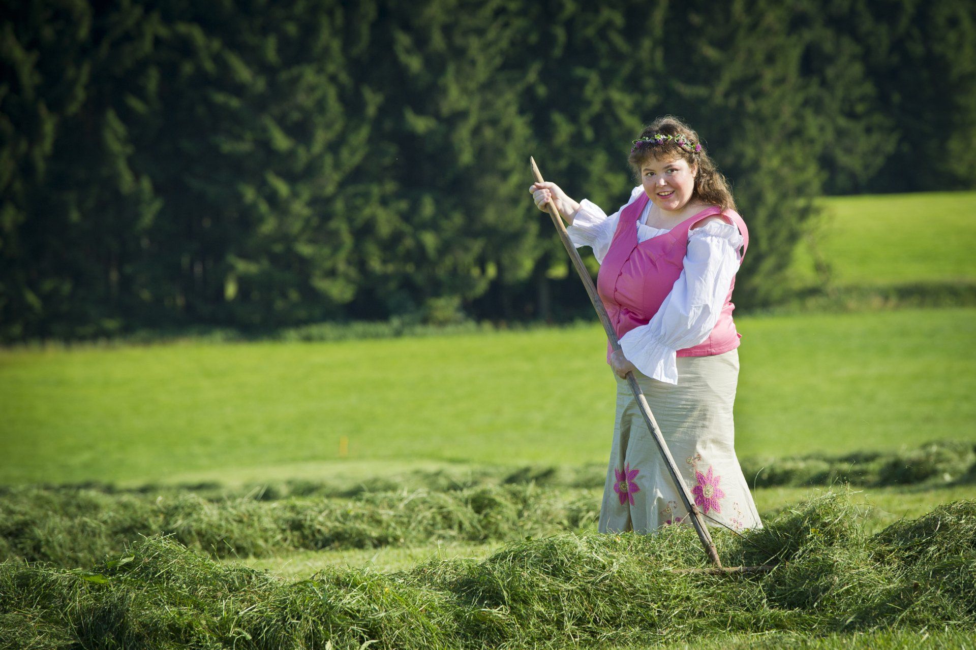 Auf dem Bild ist eine junge Frau auf einem Feld zu sehen. Sie hat eine Harke in der Hand und wendet das gemähte Gras.