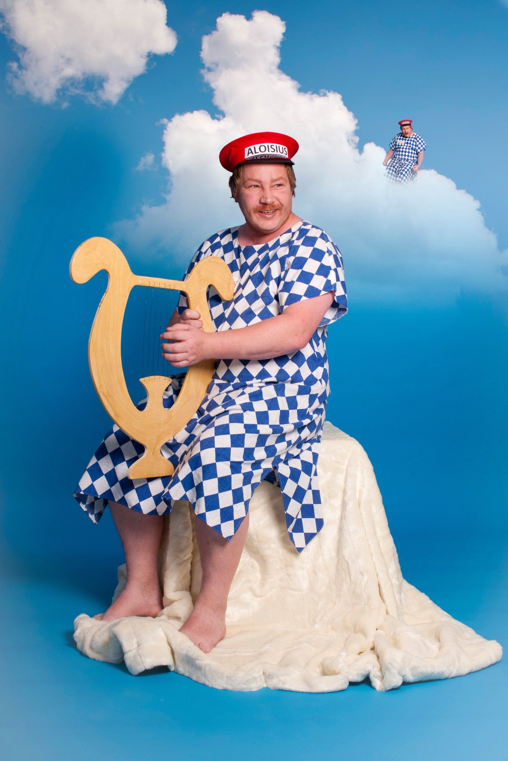 Auf diesem Bild ist Herr Freudenberg als Aloisius aus einer bekannten Münchener Geschichte dargestellt. Er hält eine Harfe in der Hand und sitzt auf einer als Wolke dargestelltem Hocker. Zudem trägt er eine rote Kappe mit der Aufschrift Aloisius und ein Gewand, welches das Muster der bayerischen Flagge hat.