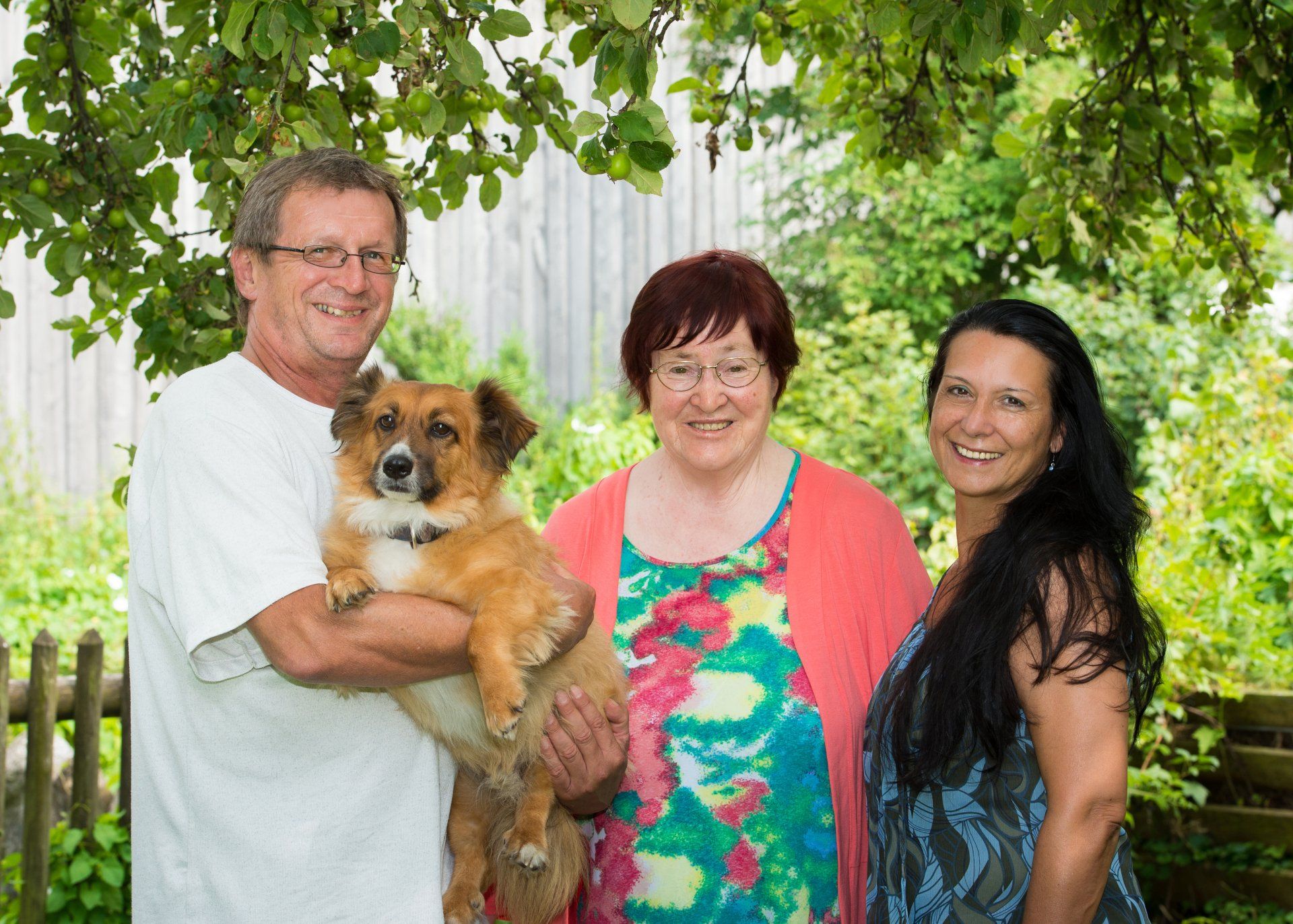 Auf diesem Foto ist Johanna mit Familie und Hund zu sehen. Alle drei stehen dicht aneinander bei einem sonnigen Tag in einem Garten.