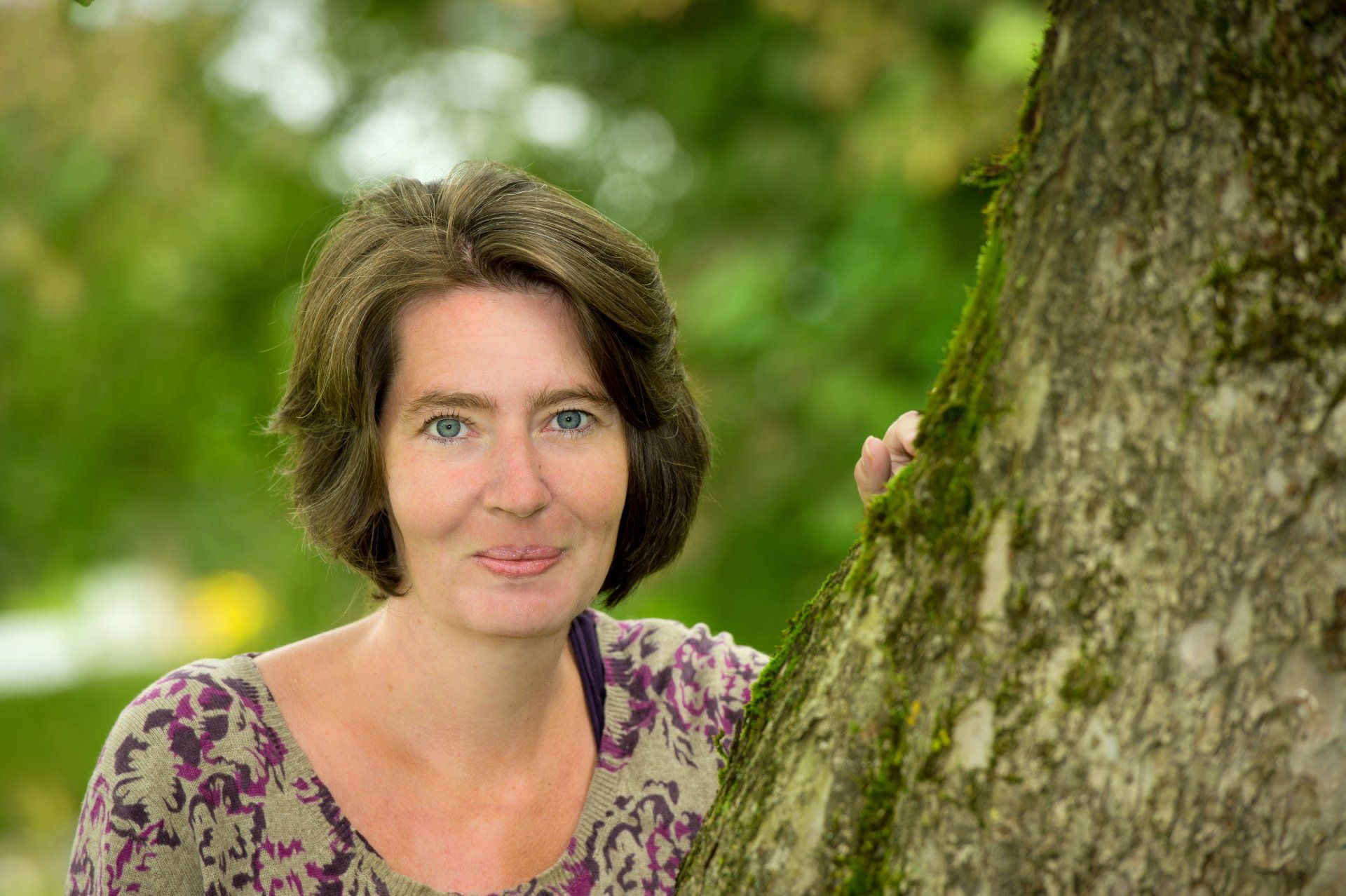 Auf diesem Bild ist Karin Jüde zu sehen, die sich für dieses besondere Foto besonders hübsch gemacht hat. Sie ist frisch frisiert, trägt Wimperntusche und ein wenig Lippenstift. Sie ist auf dem Bild in Nahaufnahme zu sehen und posiert neben einem Baum in der Natur.