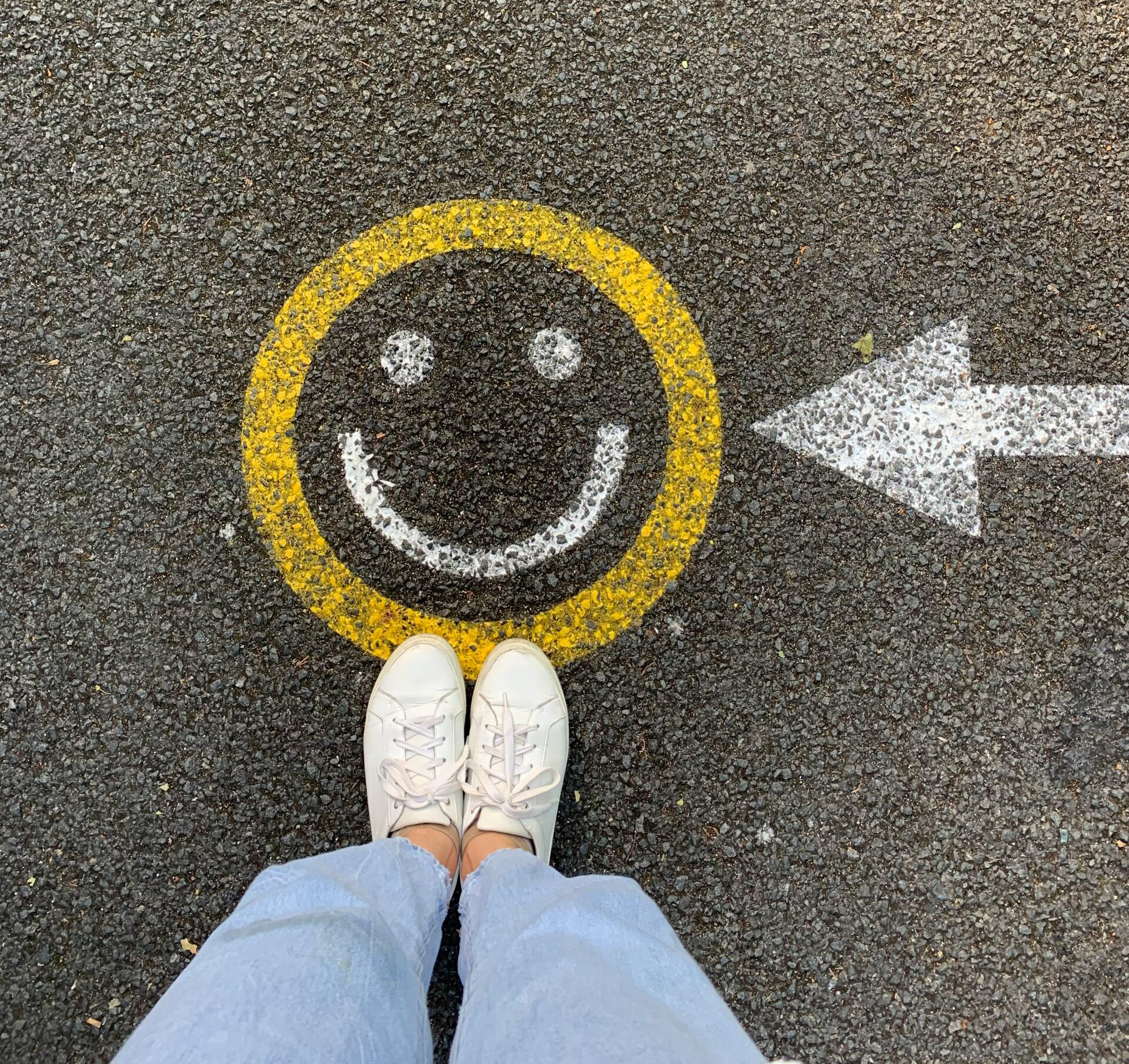 Dieses Bild ist von oben herab fotografiert. Es ist ein lächelnder Smiley zu sehen, der auf die Straße gemalt wurde. Dort ist auch ein Pfeil gemalt, der nach links auf den Smiley zeigt. Vor dem Smiley steht eine Person, deren Beine zu sehen sind.