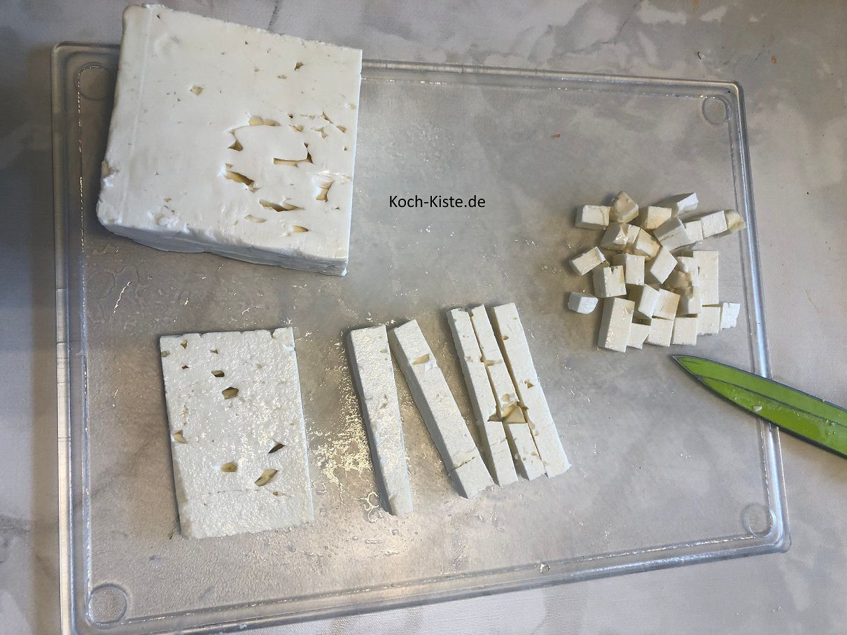 Als nächstes lege den Feta auf ein Schneidebrett und schneide ihn zuerst in Scheiben, dann in Streifen und zum Schluss in kleine Teile.