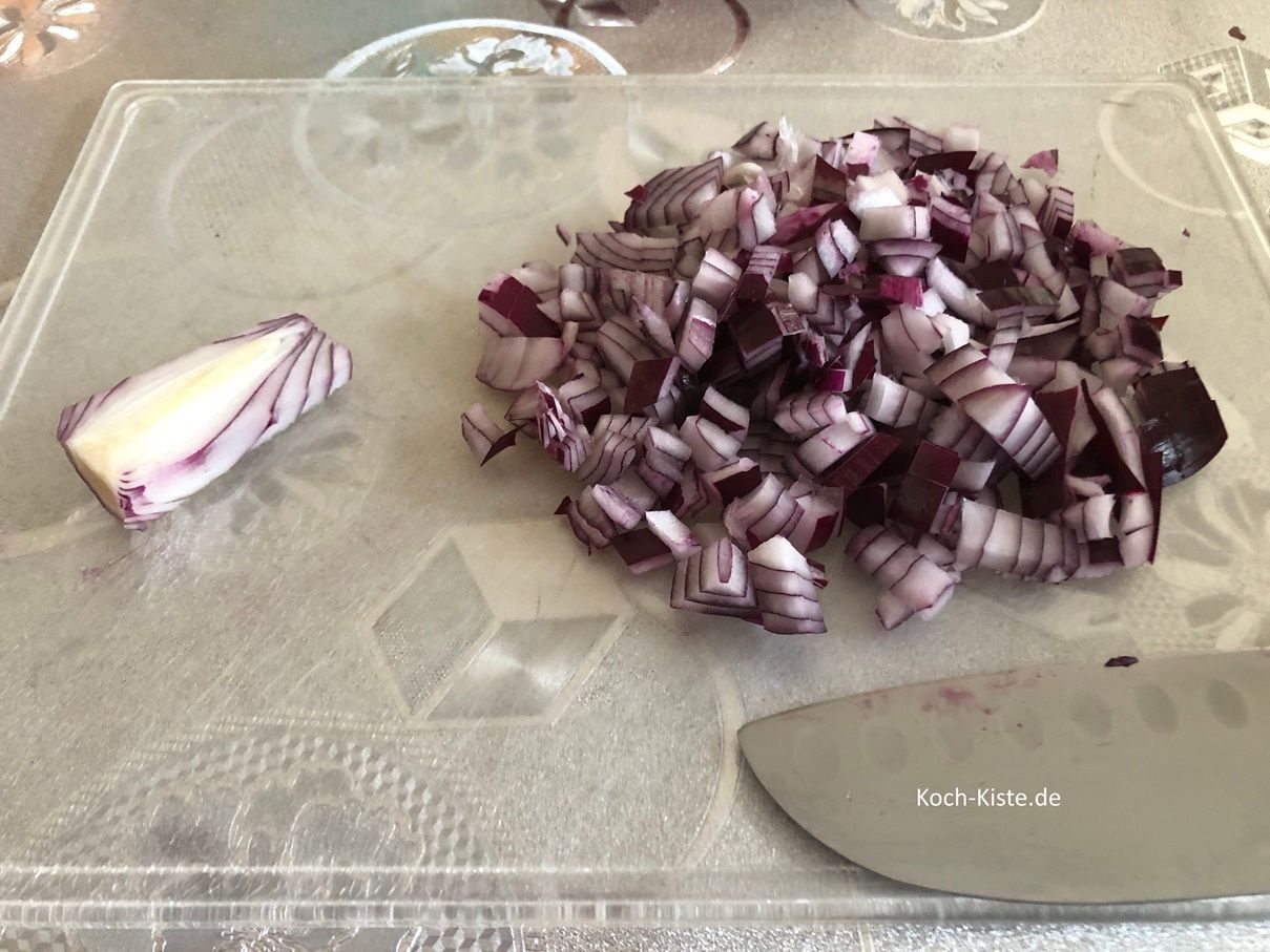 beginne damit die Zwiebel auf das Schneidebrett zu legen, oben und unten eine kleine Scheibe abzuschneiden und dann die Zwiebel zu pellen. Danach legst Du die Zwiebel wieder auf das Schneidebrett und schneidest sie erst in Scheiben, dann in Streifen und zum Schuss in kleine Stücke.