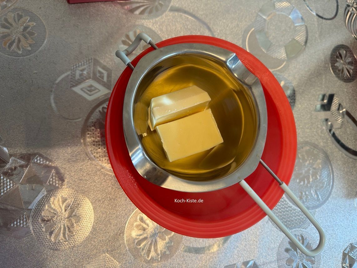 lege die Schmelzschale auf eine passende Schüssel, gebe 100 g weiße Kuvertüre in die Schmelzschale und gebe in die untere Schüssel kochendes Wasser. Dann rühre mit einem Teelöffel die Kuvertüre so lange bis sie flüssig geworden ist.