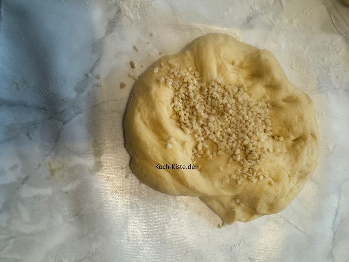 In der Zwischenzeit stelle eine weitere Schale auf die Küchenwaage auf 0 und wiege Dir 60 g gehackte Mandeln ab. Nehme eine Teigportion , drücke sie etwas flach und schütte die Mandeln hinauf. Nun musst Du die Mandeln in den Teig einkneten .