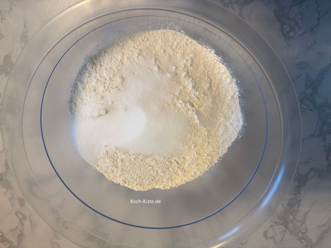 stelle die große Rührschüssel auf die Küchenwaage auf 0 und wiege 750g Dinkelmehl hinein. Danach stellst Du die kleine Schüssel auf die Küchenwaage auf 0 und wiegst 105 g Zucker ab. Schütte den Zucker mit in die große Rührschüssel.