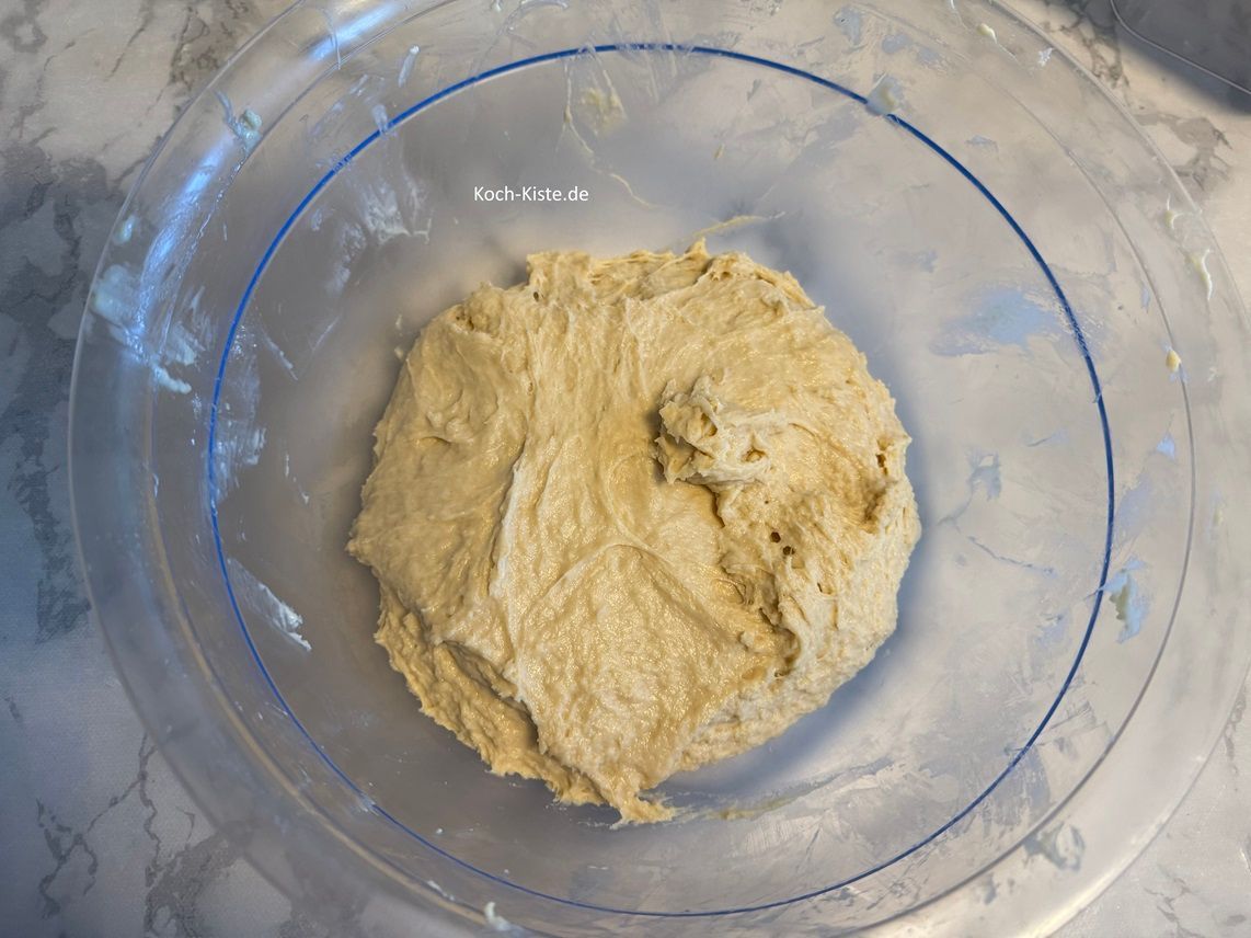 jetzt gieße die Hefemilch mit in die Rührschüssel . Ziehe Dir Vinyl-Einmalhandschuhe an und verknete die Zutaten zu einem glatten Teig. Mit einem Teigspachtel wird der Teighaufen etwas zusammen geschoben.