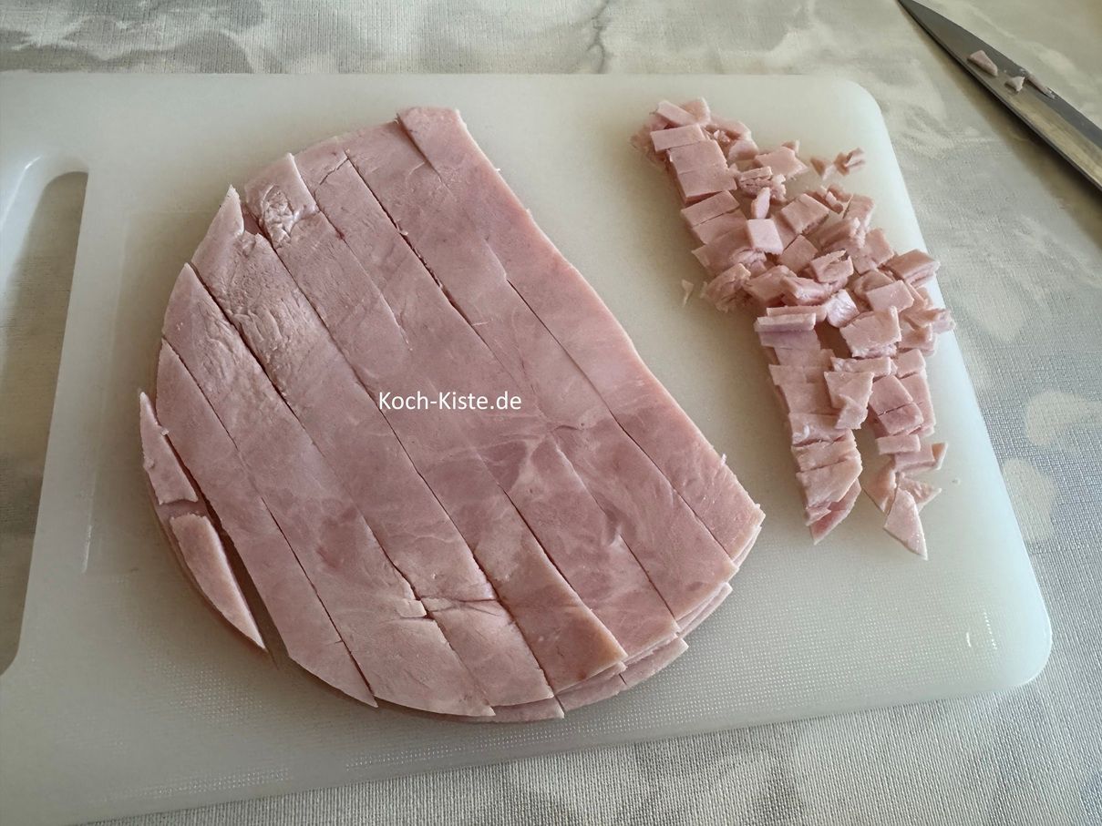 Als nächstes nehme den gekochten Schinken aus der Verpackung und lege ihn auf das Schneidebrett. Schneide den gekochten Schinken erst in schmale Streifen und dann in kleine Stücke. Zum Schluss gebe die Schinkenstücke in eine Rührschüssel.