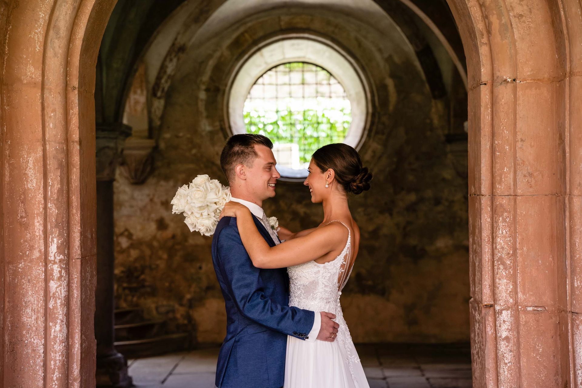 Brautpaarshooting Kloster Bronnbach im Kreuzgang, Brautpaar steht unter einem Bogendurchgang und schauen sich an.