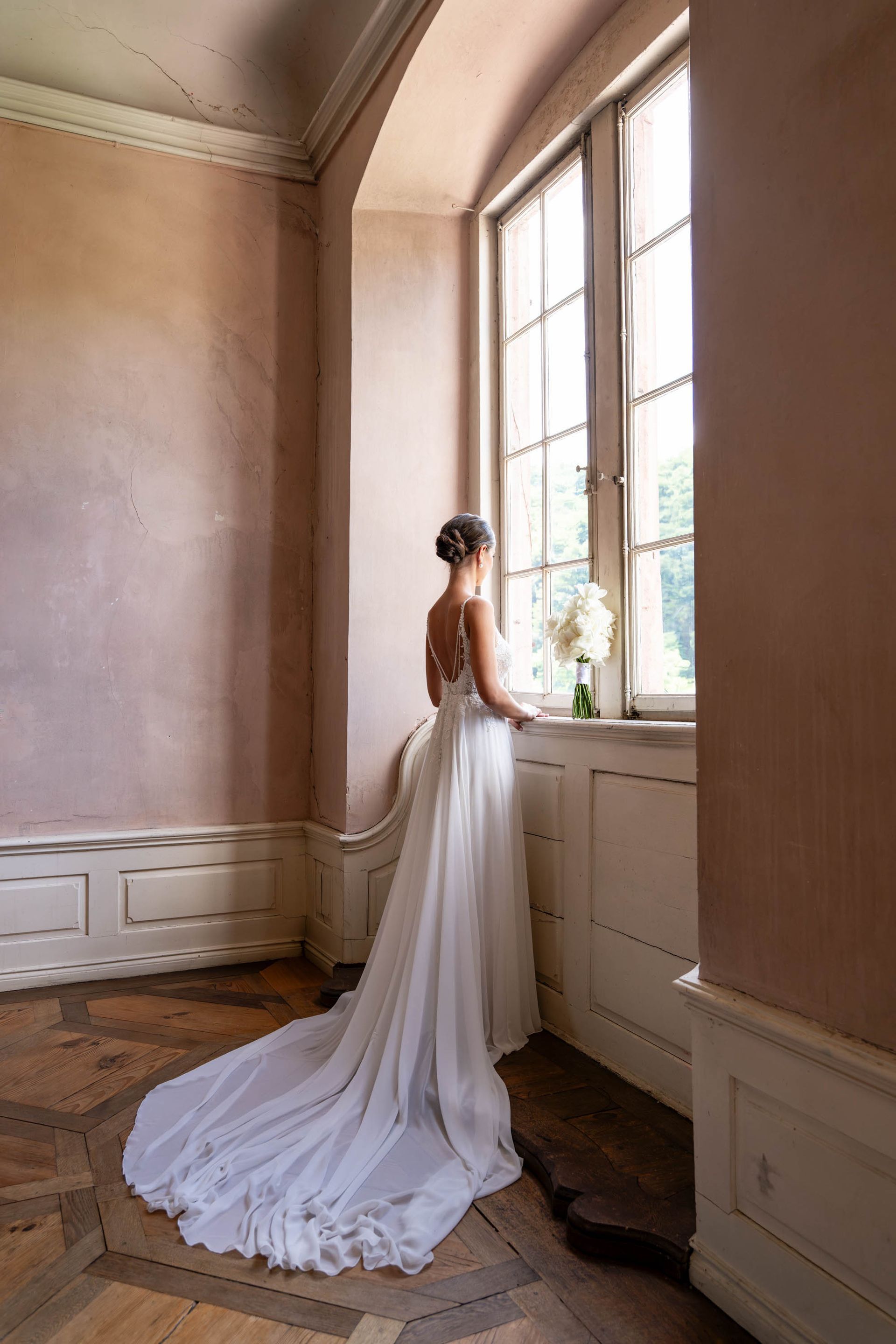 Brautpaar steht vor einer großen Fensterfront, der Bräutigam hält die Braut von hinten.