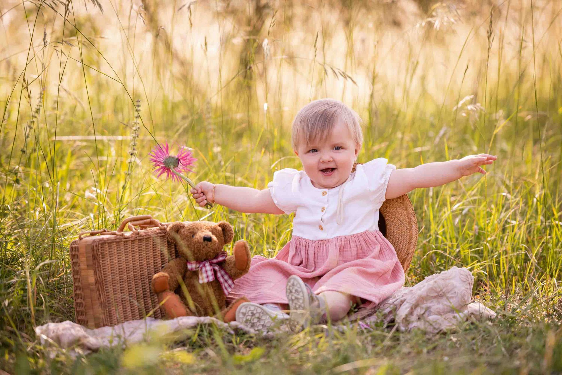 Lachendes Kleinkind in rosa Kleid sitzt auf einer Wiese mit Teddy und Korb