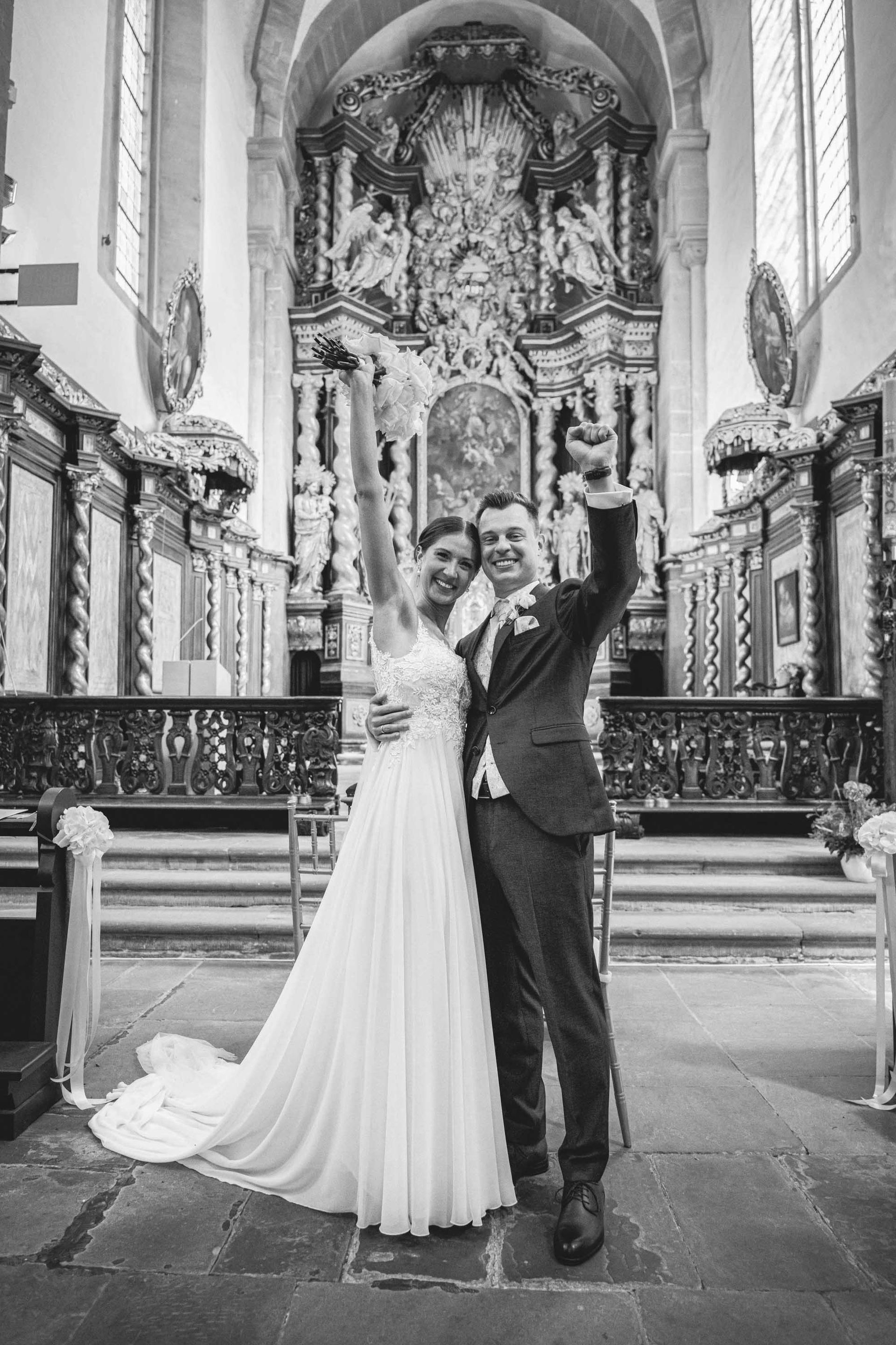 Schwarz-weiß Foto. Klosterkirche im Kloster Bronnbach kirchliche Trauung, Brautpaar steht vor dem Altar. Braut und Bräutigam schauen in die Kamera und jubeln.