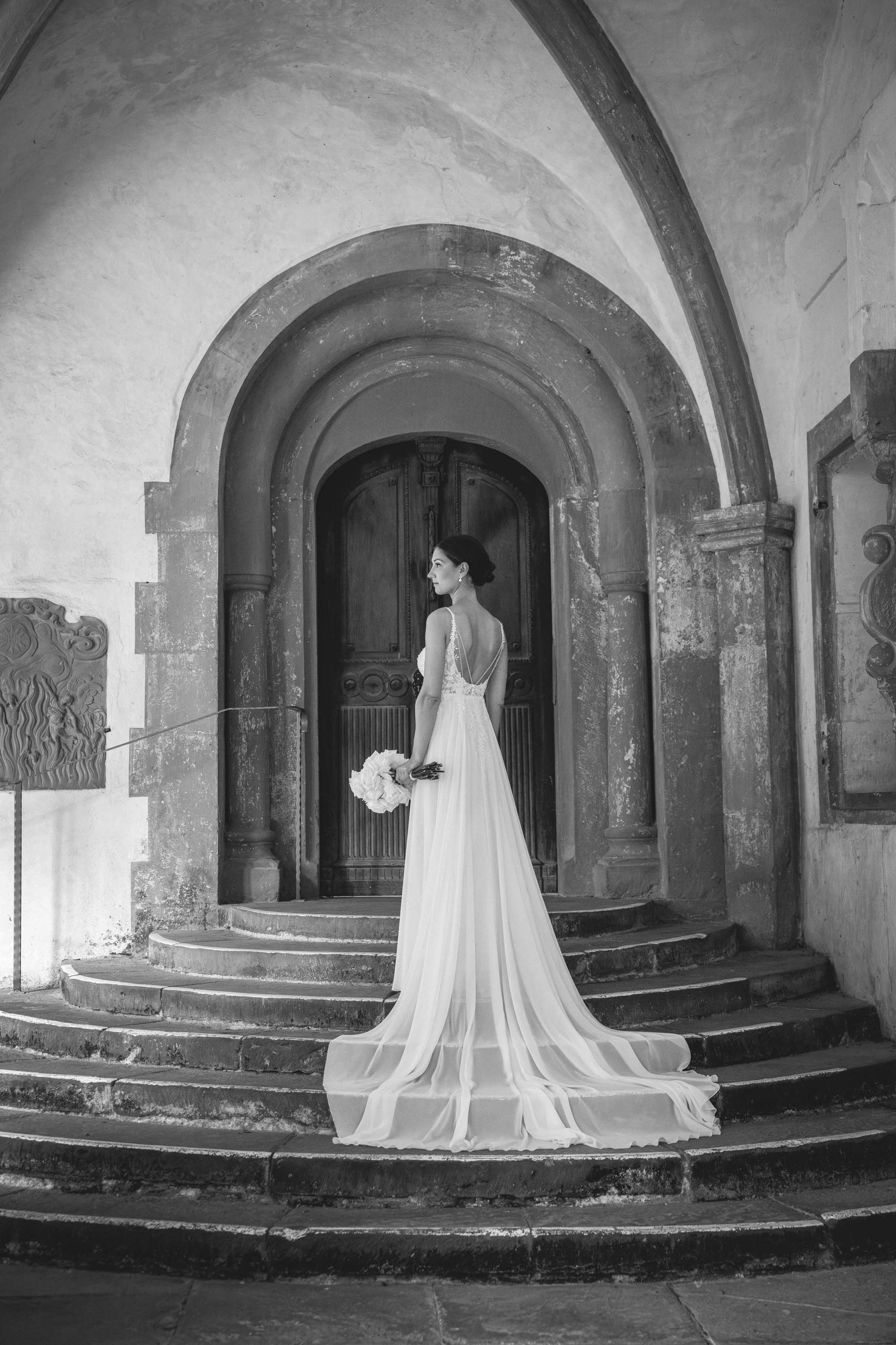 Schwarz-weiß Foto Brautpaarshooting Kloster Bronnbach im Kreuzgang. Braut steht auf Treppe. Schleppe liegt ausgebreitet auf den Treppenstufen.