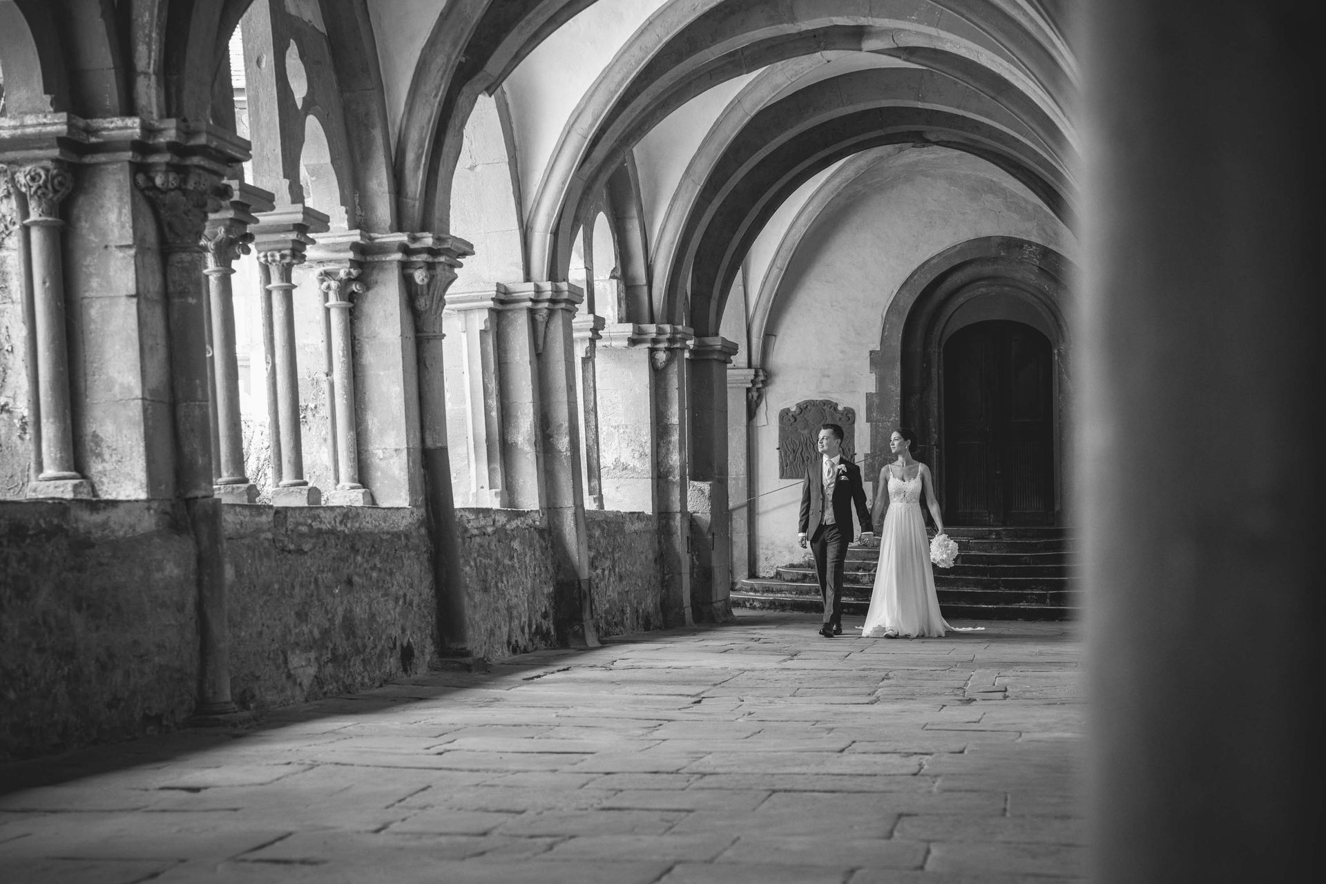 Schwarz-weiß Foto Brautpaarshooting Kloster Bronnbach im Kreuzgang. Brautpaar laufen und schauen nach vorne.