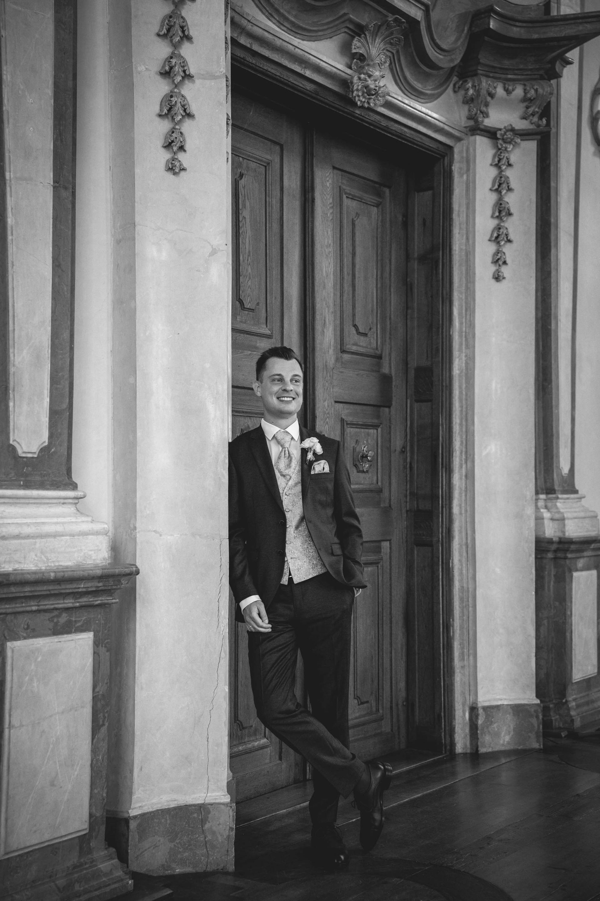 Brautpaarshooting Kloster Bronnbach im Josephsaal, mit Bräutigam im Anzug lehnt an antiker Tür.