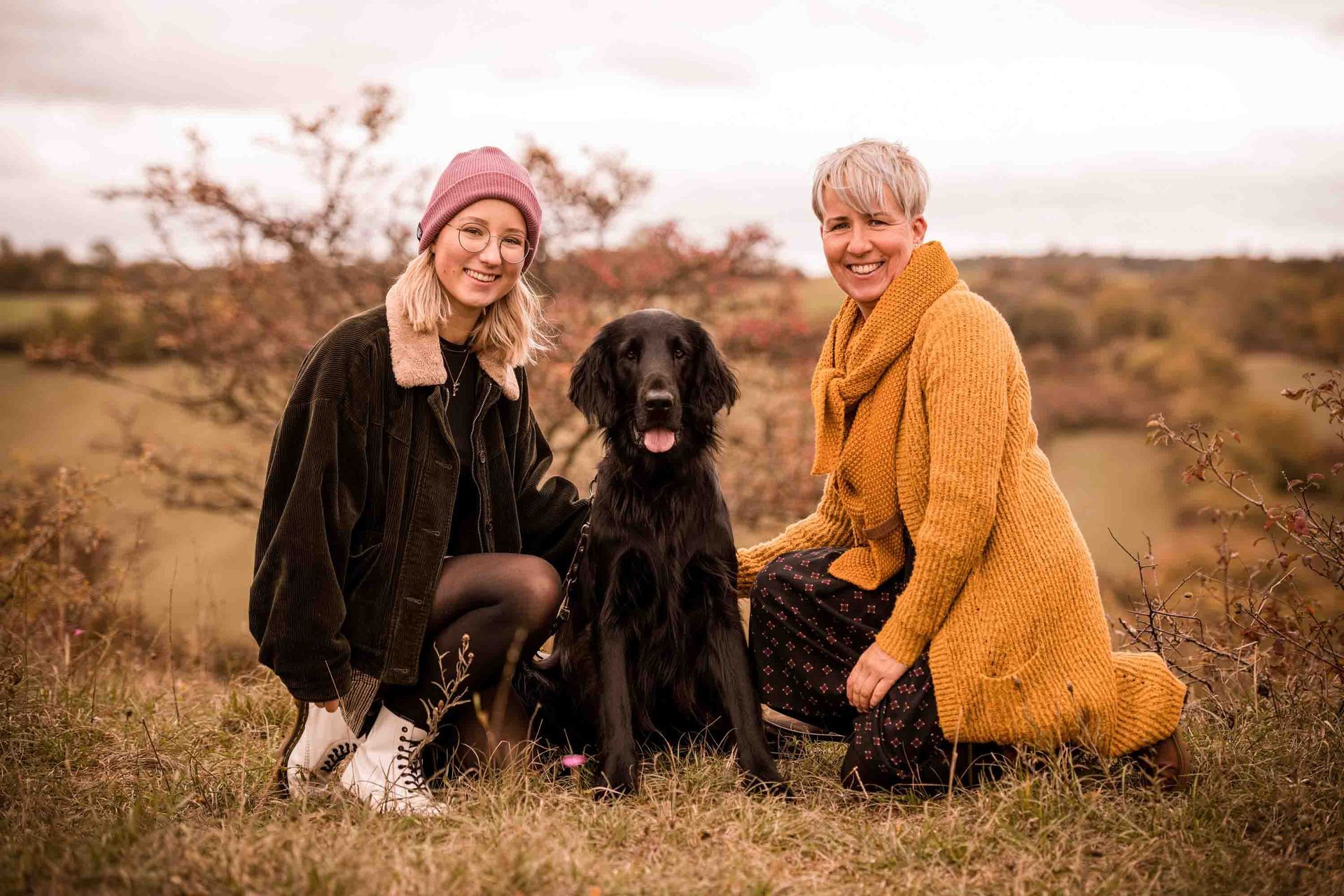 Zwei Frauen sitzen mit einem schwarzen Hund in einer Landschaft